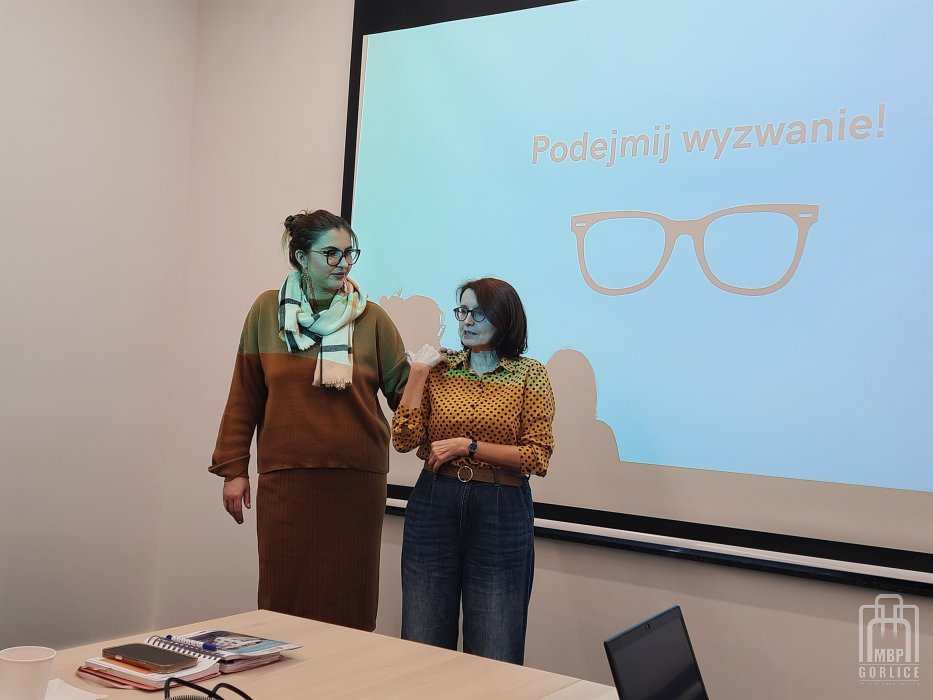 Biblioteka Gorlice, Szkolenie o odstępności ćwiczenie – prowadząca stoi wraz z uczestniczką na tle ekranu i tłumaczy jak asystować osobie niewidomej